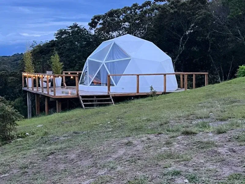 Domo geodésico blanco sobre deck de madera en ladera verde, rodeado de bosque, iluminado al atardecer