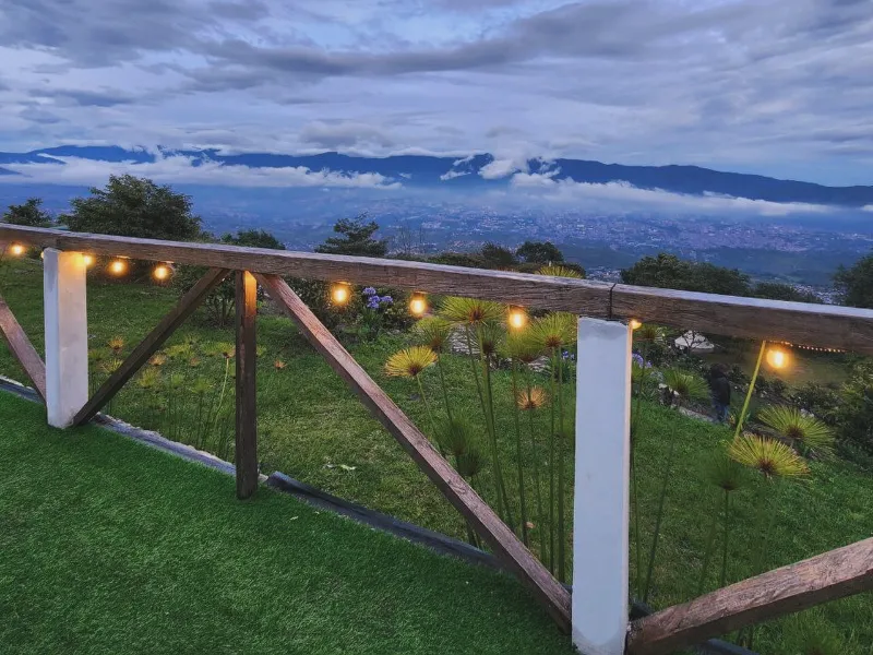 Jardín con grama, plantas silvestres y guirnalda de luces cálidas, con vista panorámica al valle y montañas entre nubes