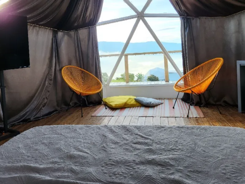 Vista desde la cama del domo hacia dos sillas naranjas tipo Acapulco frente al ventanal geodésico con paisaje montañoso