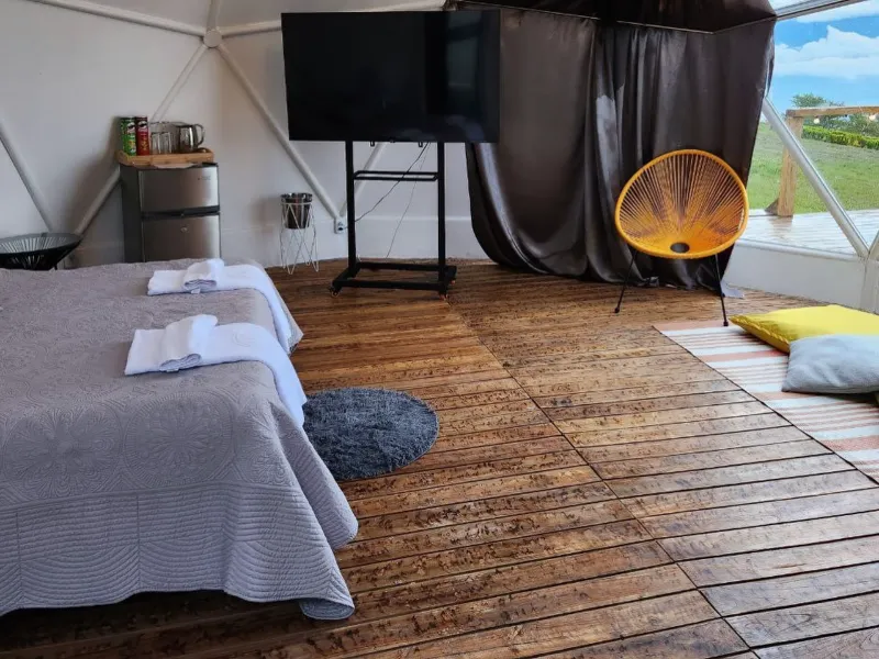 Interior de domo con cama doble, televisor en pedestal, minibar, silla naranja tipo Acapulco y piso de madera rústica