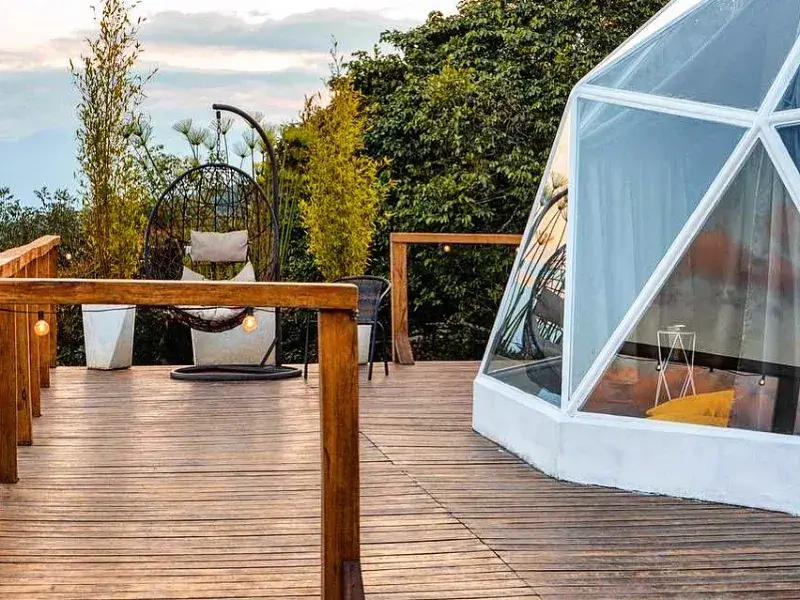 Deck de madera con mesa, silla colgante tipo huevo, fogón y jardín, junto a la estructura transparente del domo al atardecer
