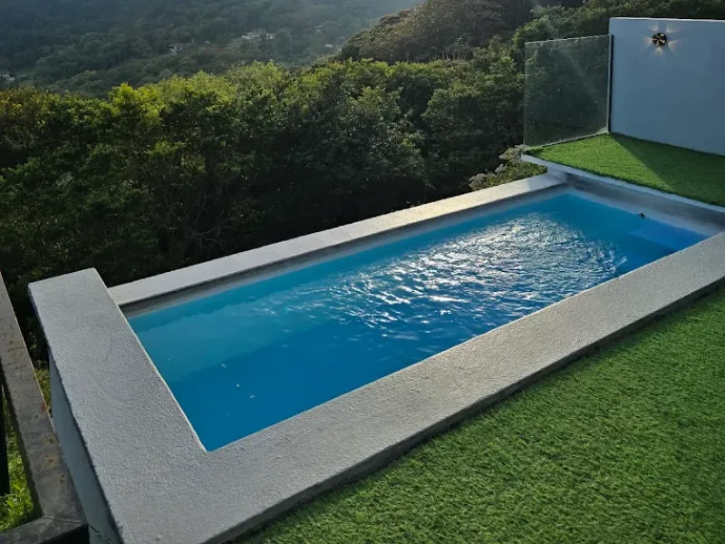 Piscina rectangular de agua azul rodeada de grama artificial, con vista al bosque y montañas verdes