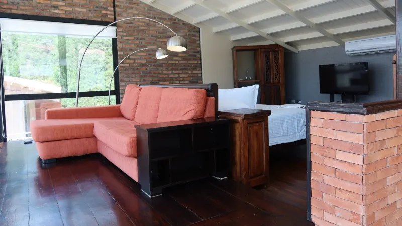 Sala-habitación con sofá naranja en L, piso de madera oscura, pared de ladrillo visto y cama doble al fondo con televisor