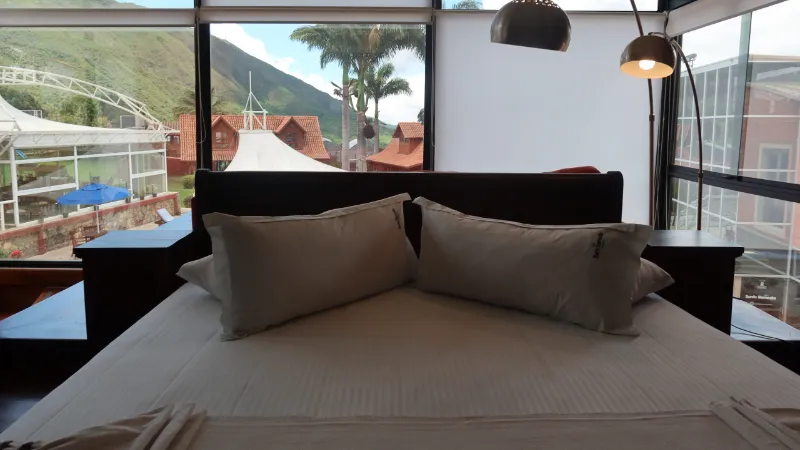 Cama doble con ropa blanca y lámpara de arco, frente a ventanal panorámico con vista a cabañas, piscina, palmeras y montaña