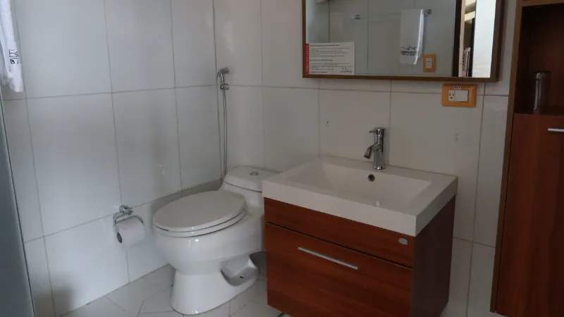 Baño privado con sanitario, lavamanos empotrado en mueble de madera, espejo y paredes de cerámica blanca