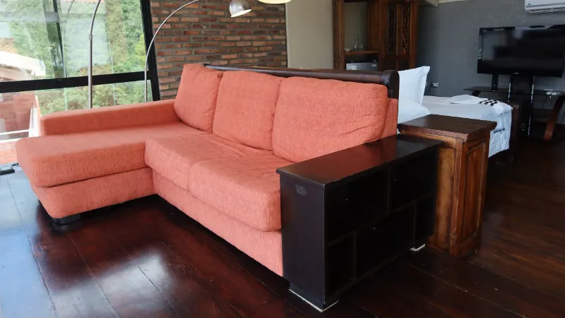 Sofá esquinero color naranja sobre piso de madera oscura, con ventanal al jardín, lámpara de arco y zona de descanso al fondo