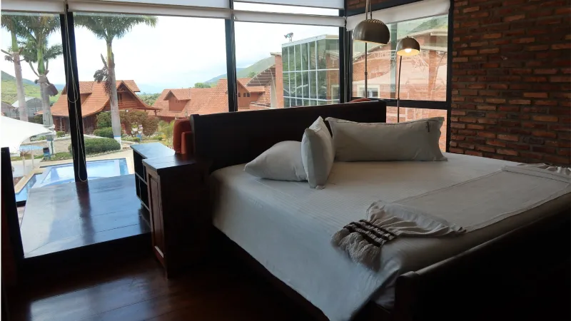 Cama king con ropa blanca y pared de ladrillo visto, junto a ventanal con vista a piscina, cabañas y montañas verdes