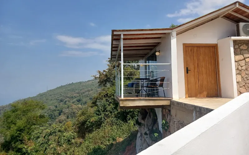 Cabaña moderna con terraza voladiza, barandal de vidrio y cable, puerta de madera y aire acondicionado, con vista a ladera boscosa y mar al fondo bajo cielo azul.