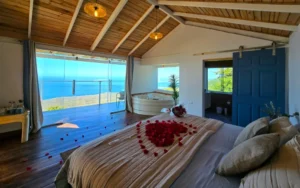 Habitación romántica con cama decorada con pétalos de rosas rojas en forma de corazón, jacuzzi, techo de madera a dos aguas, puerta corrediza azul y ventanal con vista al mar en Villa Amalfi LaVista Glamping