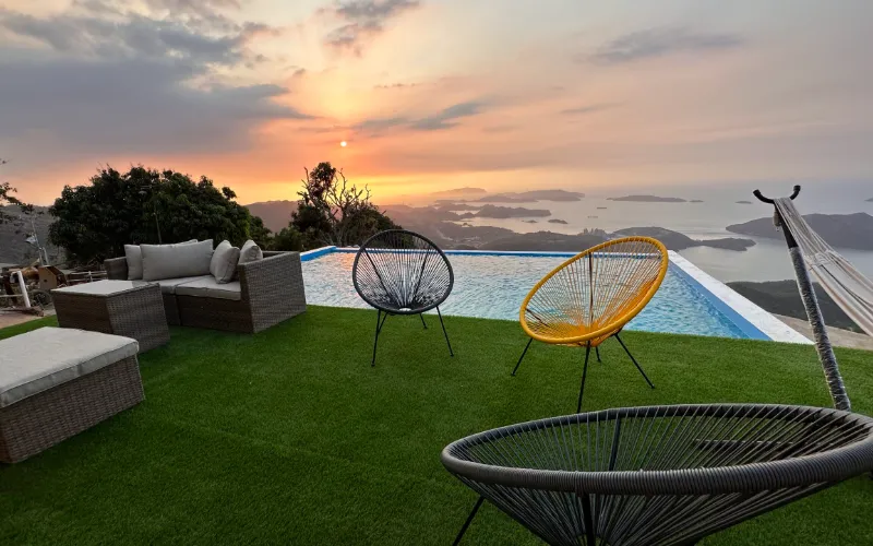 Área social con sillas tipo acapulco negra y amarilla, sofá de ratán, hamaca y piscina infinita al atardecer, con archipiélago de islas y cielo anaranjado al fondo.