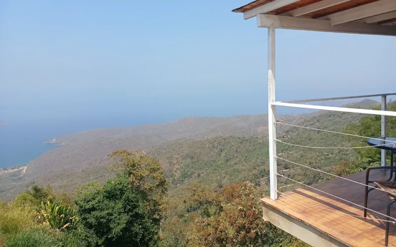 Terraza con barandal de cable metálico y silla, con vista panorámica a laderas boscosas, costa y mar azul bajo cielo despejado.
