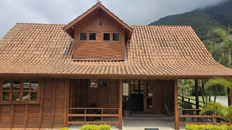 Fachada de cabaña de madera con techo de teja a dos aguas y buhardilla, terraza cubierta con barandas y montaña al fondo
