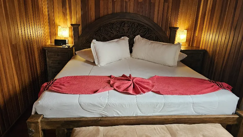 Cama king con cabecera tallada en madera oscura, ropa blanca con detalle rojo en lazo, flanqueada por lámparas de noche cálidas