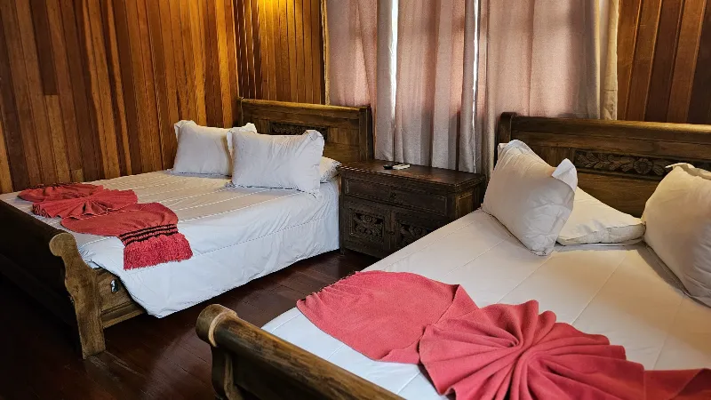 Habitación con dos camas individuales de madera tallada, ropa blanca con toallas rojas decorativas y paredes de madera oscura