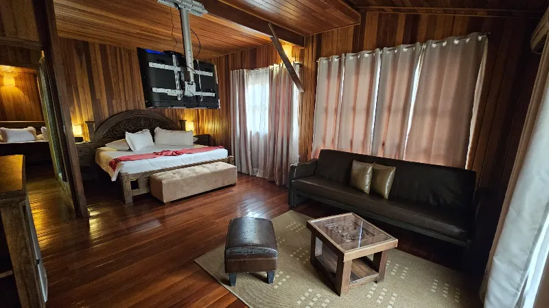 Suite con cama king, sala con sofá de cuero negro, televisor de techo giratorio y piso de madera en cabaña rústica