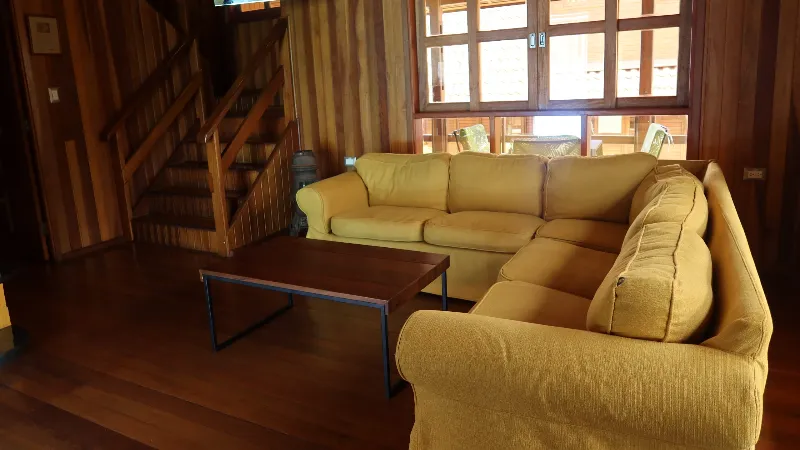 Sala de estar con sofá esquinero amarillo, mesa de centro, escalera de madera y ventana panorámica en cabaña de dos pisos