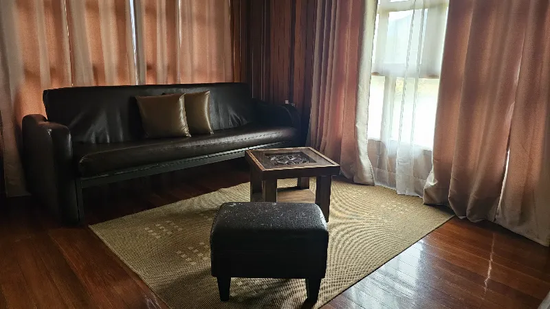 Sala íntima con sofá de cuero negro, puf, mesa de centro tallada y cortinas rosas sobre piso de madera oscura