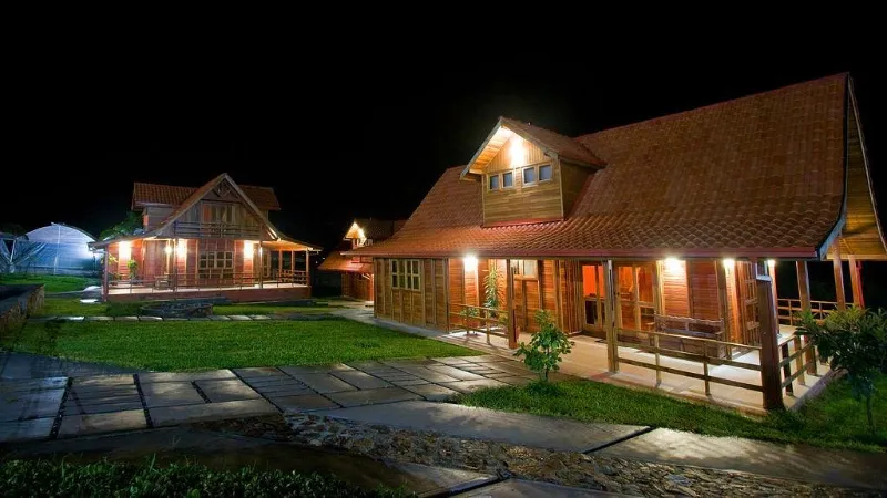 Vista nocturna de dos cabañas de madera con techo a dos aguas iluminadas cálidamente, rodeadas de jardín verde con sendero de piedra