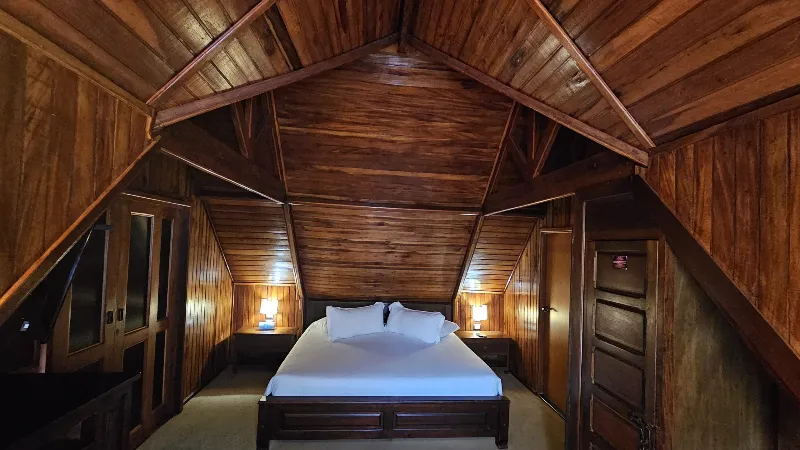 Habitación tipo loft en cabaña con techo triangular de madera, cama doble blanca y lámparas de noche a ambos lados