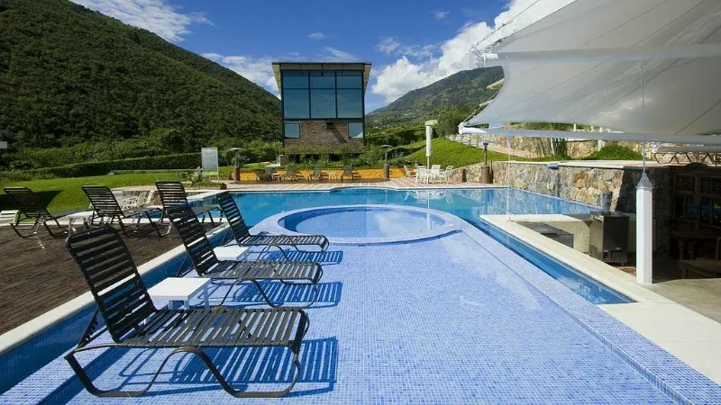 Piscina azul con jacuzzi integrado, sillas de playa, edificio de ladrillo y vidrio al fondo, rodeada de montañas verdes