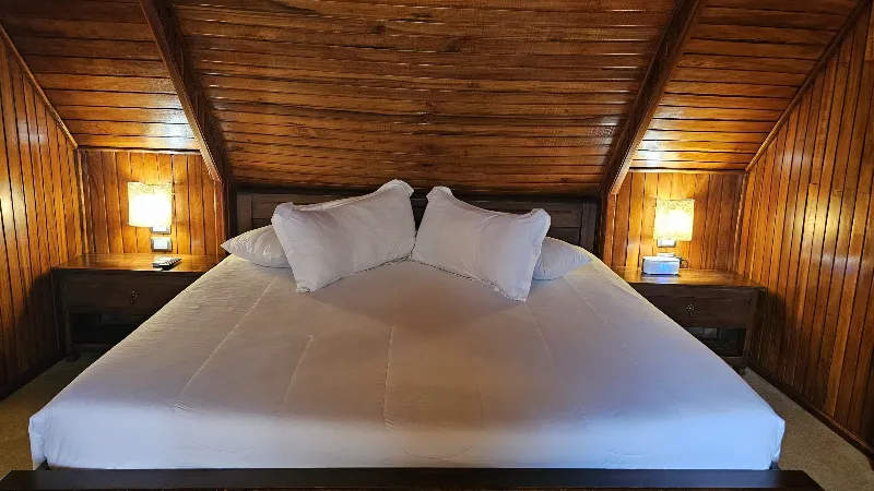 Cama king con ropa blanca y dos lámparas de noche, enmarcada por techo y paredes inclinadas de madera oscura