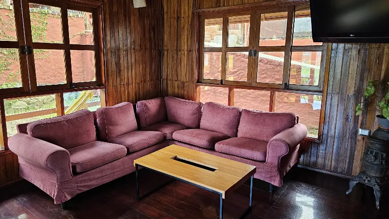 Sala de estar con sofá esquinero vino tinto, mesa de centro de madera, televisor y ventanas con vista al exterior en cabaña de madera