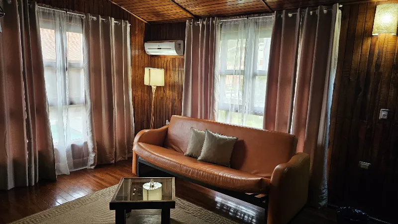 Sala acogedora con sofá de cuero camel, cortinas rosadas, lámpara de pie y aire acondicionado en cabaña de madera