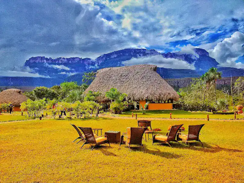 Área de descanso del campamento con sillas de ratán sobre pradera dorada, churuata central con techo de palma y tepuyes majestuosos entre nubes al fondo.