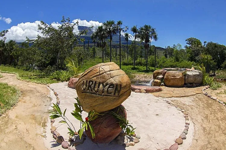 Roca de bienvenida con el nombre "Uruyen" grabado, fuente de agua natural, palmeras moriche y tepuy bajo cielo azul despejado.