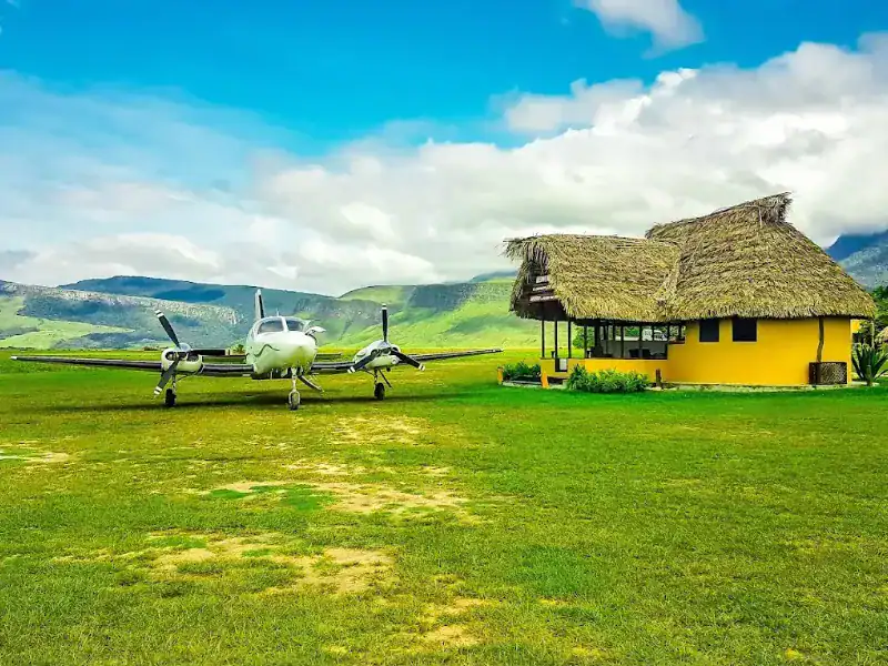 Avioneta bimotor estacionada junto a churuata amarilla de techo de palma en la pista de aterrizaje del campamento, con tepuyes verdes al fondo.