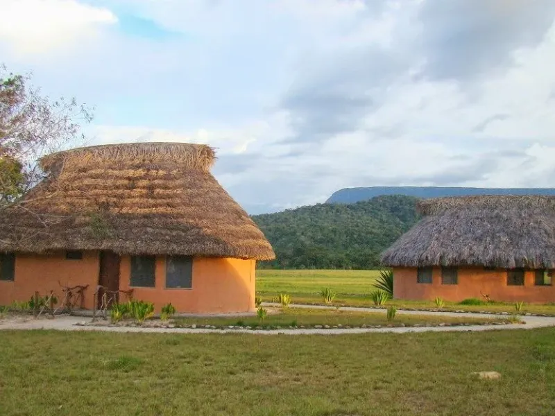 Dos churuatas circulares de paredes naranjas y techo de palma al atardecer, rodeadas de sabana verde y tepuy al fondo bajo cielo nublado.