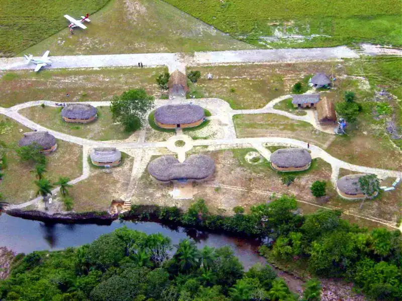 Vista aérea del Campamento Uruyen mostrando distribución de churuatas con techos de palma, senderos, pista de aterrizaje con avionetas y río bordeando el
