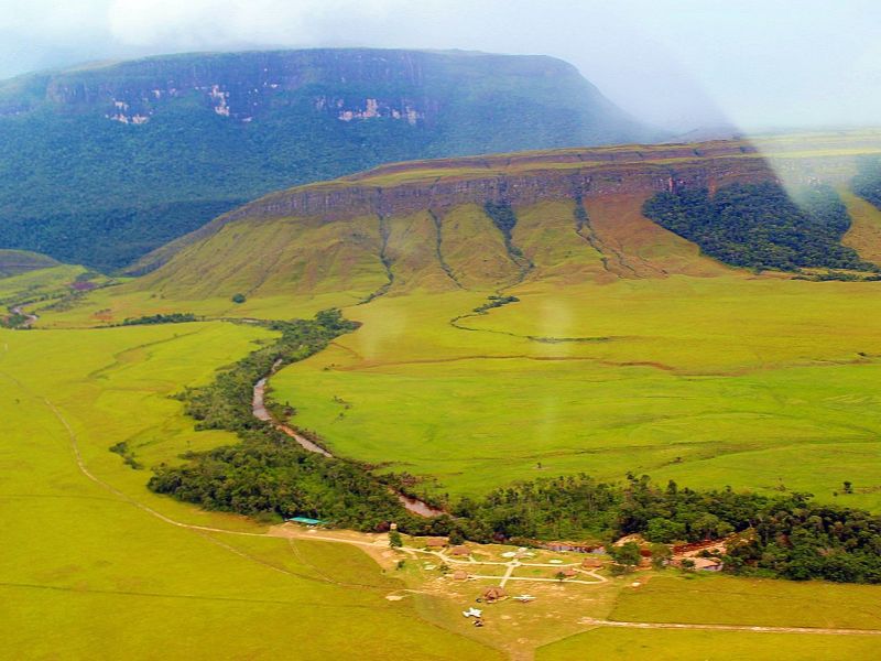 Vista aérea de la Gran Sabana con río serpenteante, tepuy de cima plana, sabana verde extensa y las instalaciones del campamento al pie de la montaña.