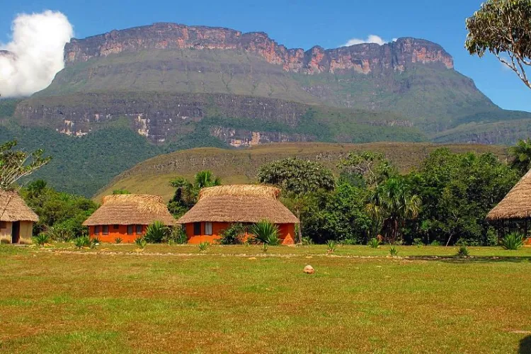 Conjunto de churuatas circulares de paredes naranjas y techo de palma sobre sabana verde, con imponente tepuy de paredes verticales bajo cielo azul.