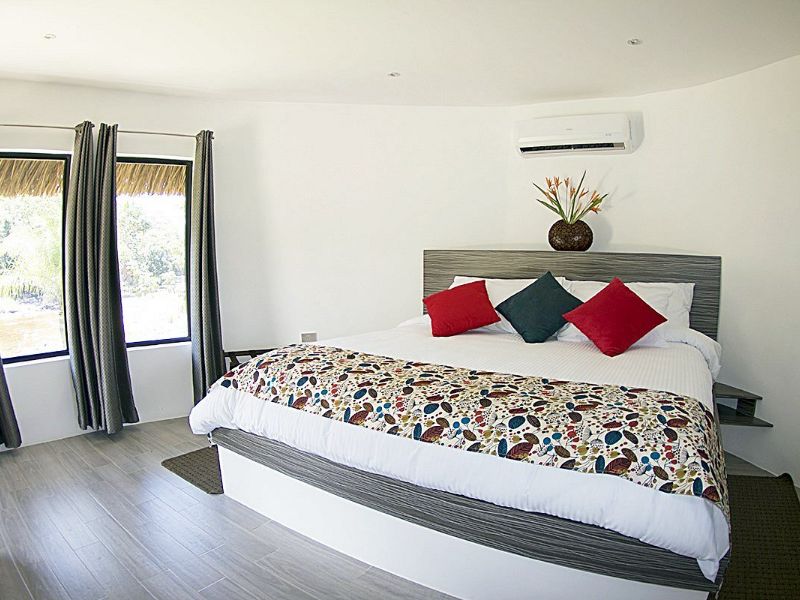 Habitación moderna con cama doble, almohadas rojas y verdes, cubrecama floral, aire acondicionado y ventana con vista al techo de palma.