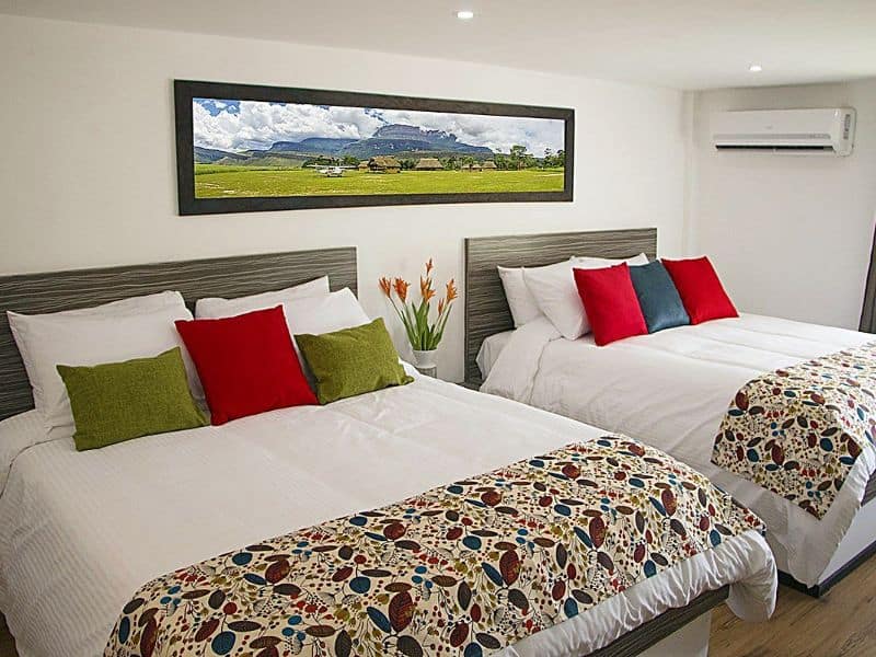 Habitación con dos camas dobles, almohadas rojas y verdes, cuadro panorámico del campamento en la pared y aire acondicionado.