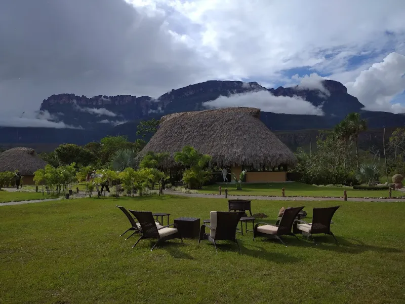 Zona social del campamento con sillas de ratán en jardín verde, churuata central y tepuyes envueltos en nubes al atardecer.
