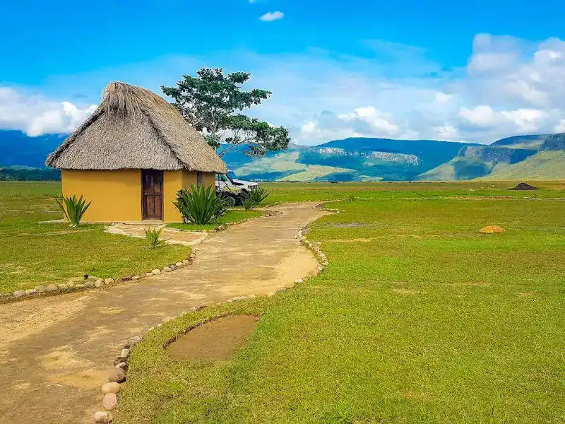 Churuata individual de paredes amarillas y techo de palma con sendero de tierra, sabana verde y tepuyes al fondo bajo cielo azul.