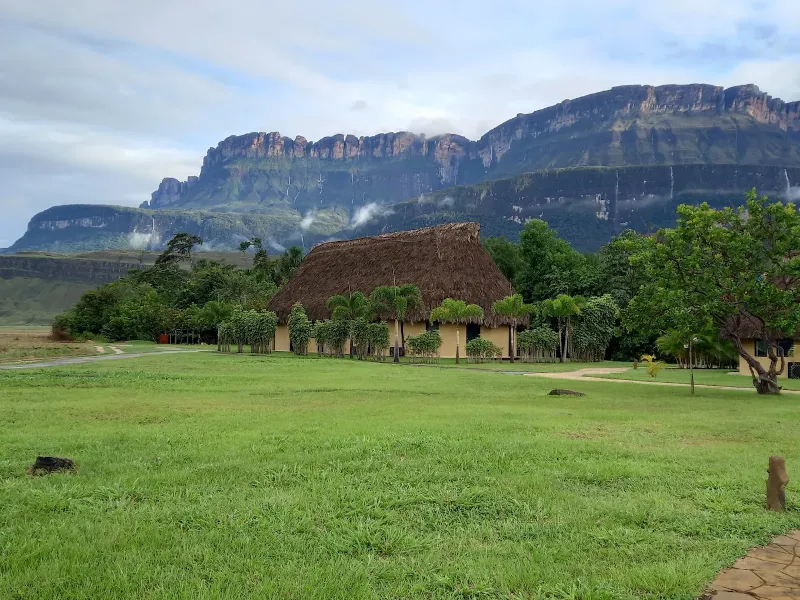 Vista del campamento con gran churuata de techo de palma, palmeras y jardín verde, con la imponente pared del Auyan-Tepui y cascadas al fondo.