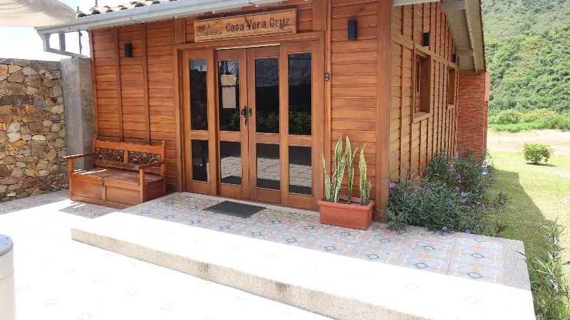 Entrada principal de Casa Vera Cruz con fachada de madera, puerta doble de vidrio, banca tallada y plantas en terraza de baldosa