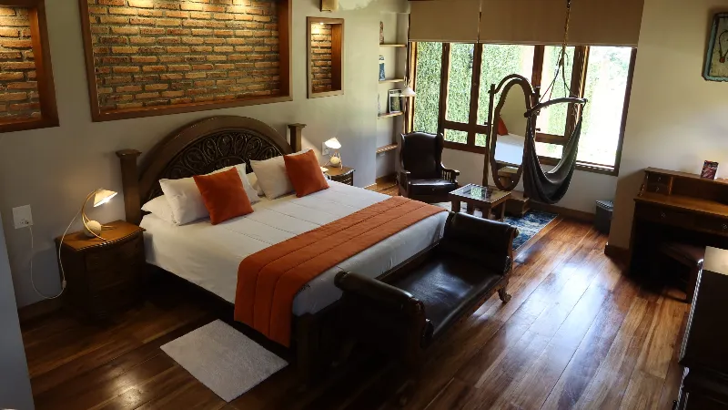 Habitación king con cabecera tallada, cojines naranjas, banco de cuero al pie, hamaca colgante y vista al jardín desde ventanas de madera