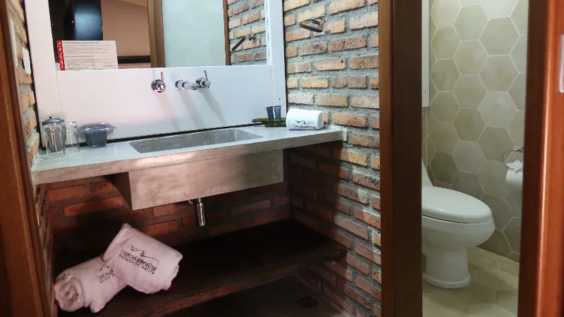 Baño con lavamanos de concreto empotrado en pared de ladrillo visto, toallas bordadas y acceso al sanitario con cerámica hexagonal