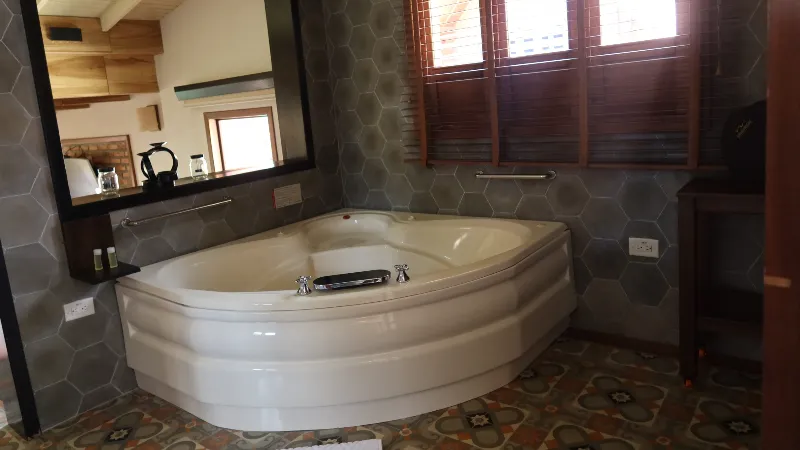 Tina de hidromasaje esquinera en baño con paredes de cerámica hexagonal gris, espejo grande y ventana de persiana de madera