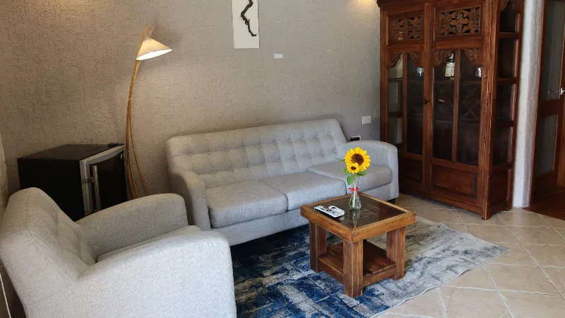 Sala con sofá y sillón grises capitoné, mesa con girasol amarillo, lámpara de pie y vitrina de madera tallada