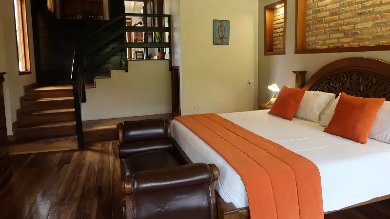 Habitación king con cojines naranjas, sillones de cuero oscuro al frente, pared de ladrillo visto y escalera de madera con barandal negro