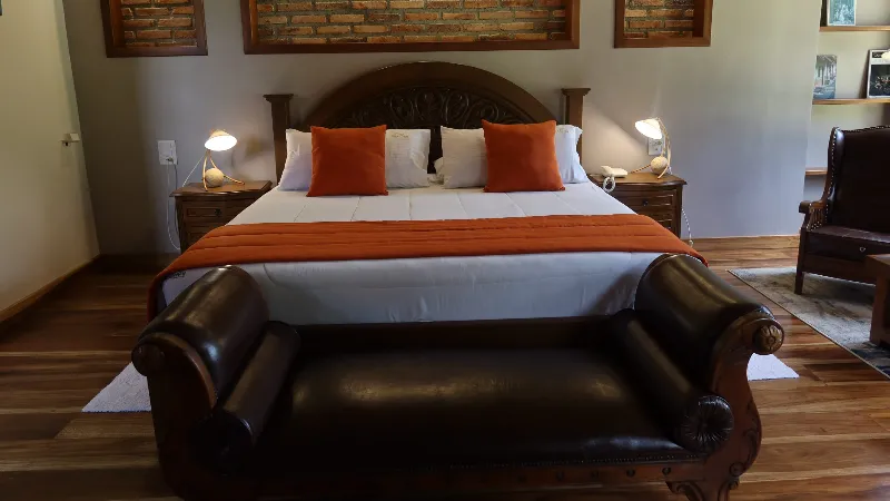 Cama king con cabecera tallada, cojines naranjas, lámparas de noche simétricas, recamier de cuero oscuro y piso de madera