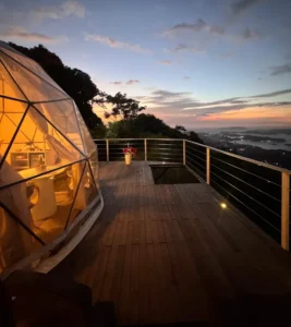 Domo geodésico iluminado en cálido tono naranja al atardecer, con deck de madera, barandal metálico y vista panorámica al mar con islas y luces urbanas al fondo.