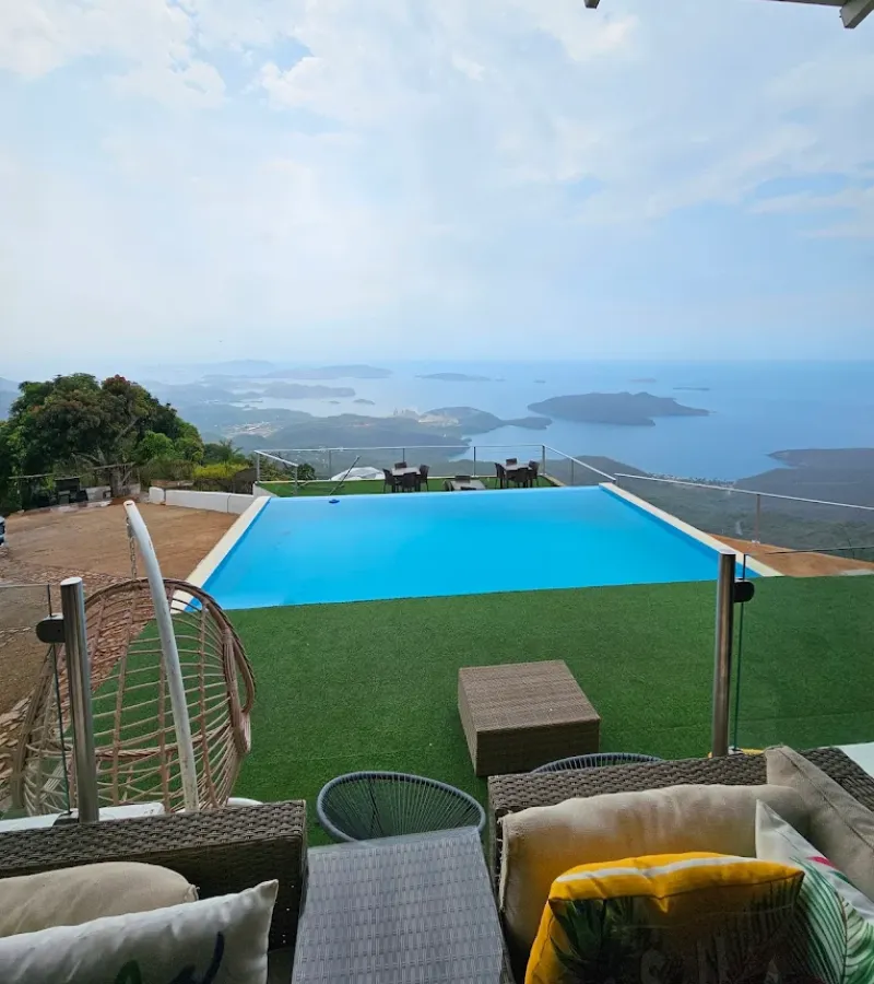 Piscina infinita azul con vista espectacular al mar y archipiélago de islas, rodeada de área de descanso con sillas de ratán, silla colgante y césped verde.
