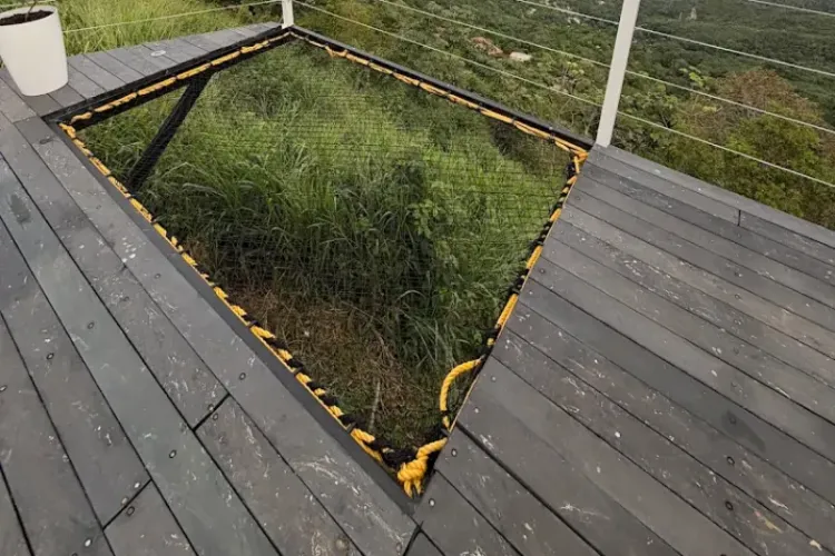 Trampolín de red sobre deck de madera con borde amarillo, con vista aérea a colinas verdes, costas y archipiélago de islas bajo cielo nublado.