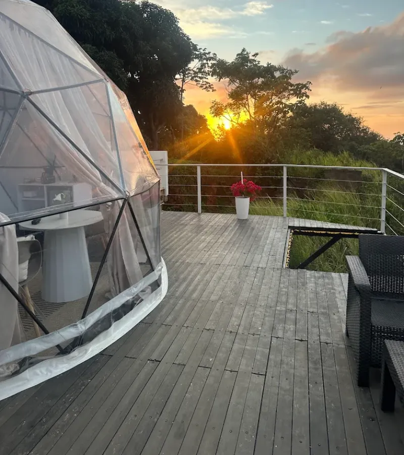 Domo geodésico en deck de madera al atardecer con rayos de sol entre árboles, maceta con flores rojas y sillas de exterior con vista a vegetación y cielo naranja.